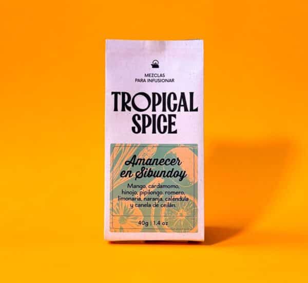Imagen del empaque biodegradable de infusión Amanecer en Sibundoy de Tropical Spice, con ingredientes como mango, cardamomo, hinojo, pipilongo, romero y caléndula. Fondo naranja intenso.