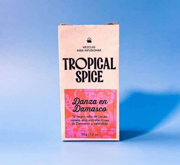 empaque biodegradable de infusión Danza en Damasco de Tropical Spice, hecha con té negro, nibs de cacao, anís estrella, canela, rosas de Damasco y caléndula. Fondo azul suave.