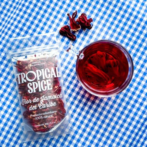 Flor de Jamaica del Caribe de Tropical Spice junto a taza con infusión roja sobre mantel de cuadros azules.