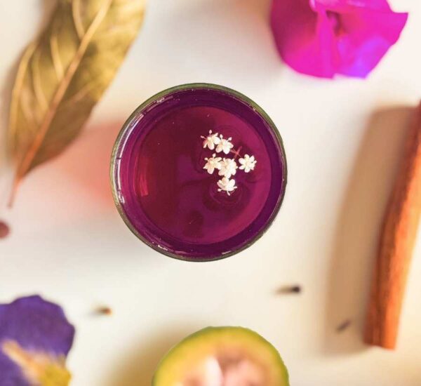 Vista superior de la bebida Amor en el Altiplano color morada decorada con flores blancas pequeñas, rodeada de ingredientes como canela, hojas secas y pétalos.