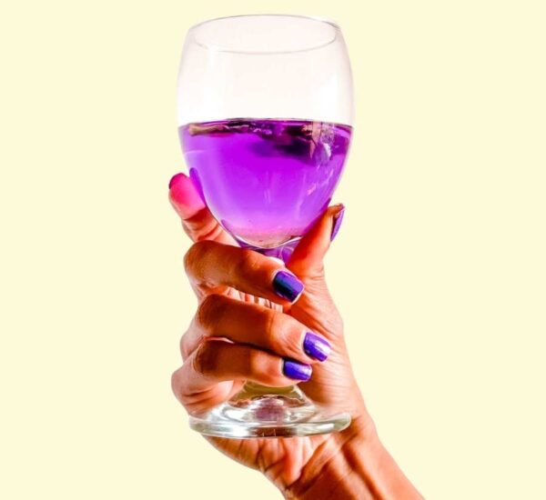 Mano con uñas moradas sostiene copa de cristal con infusión violeta de flor de clitoria.