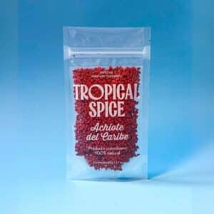 Achiote del Caribe en grano de Tropical Spice, empacado en bolsa transparente doypack sobre fondo azul degradado.