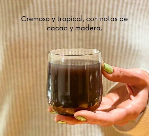 Persona sosteniendo un vaso de bebida oscura cremosa con texto que describe su sabor tropical con notas de cacao y madera.