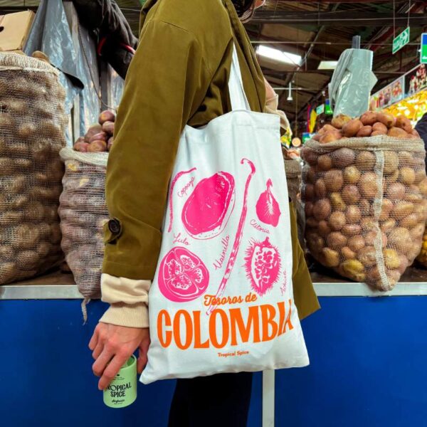 Bolsa de tela “Tesoros de Colombia” de Tropical Spice con ilustraciones en fucsia y naranja de productos como copoazú, maracuyá, clitoria y achiote, sostenida en un mercado.