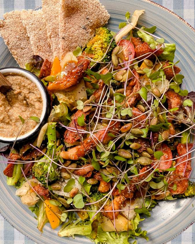 Ensalada colorida con hojas verdes, hongos salteados, mango, germinados y semillas tostadas, servida con casabe y hummus.