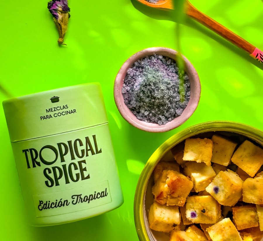 Mezcla para cocinar Edición Tropical de Tropical Spice con sal violeta y bocados horneados, sobre fondo verde brillante.